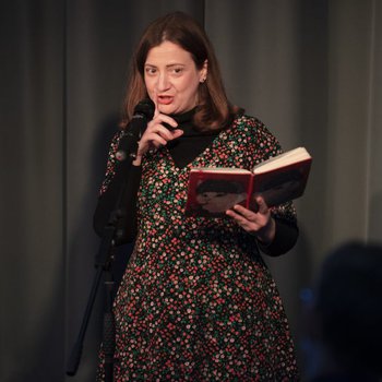 Lady on stage holding a book and reciting into a microphone