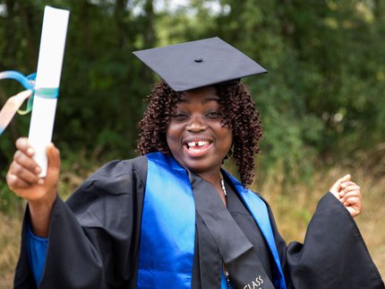 A smiling woman wearing graduation robes, holding a diploma, standing in front of trees