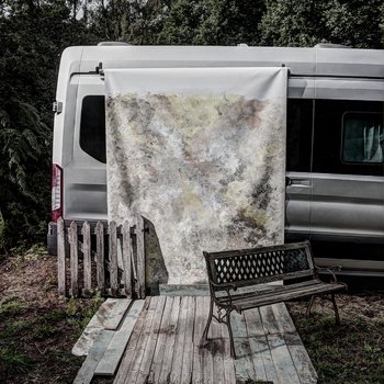 An outdoor set up for photography; a van with a textured backdrop hung down the side with pallet flooring and wooden bench in front