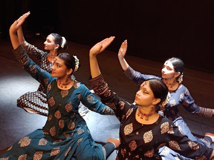 Four female dancers sit on a stage with their arms raised in a classical Indian Kathak style, wearing classical Indian dress.