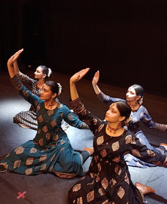Four female dancers sit on a stage with their arms raised in a classical Indian Kathak style, wearing classical Indian dress.