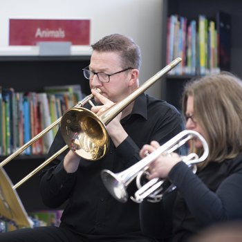 A man and a woman wearing black shirts playing brass instruments in front of shelves of books.