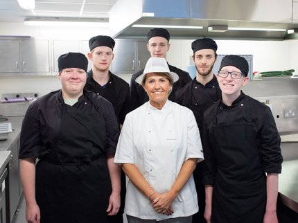 A group of Inspire College students in chef uniforms stood around a tutor in a professional kitchen.
