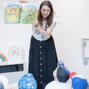 An author in a long navy skirt reads to a group of children in a school room