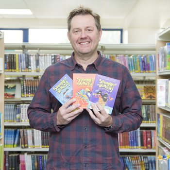 An author stands in an isle of books and holds three copies of his book