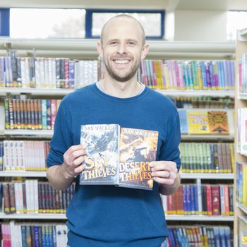 An author wearing a blue shirt stands in an isle of books and holds two copies of his book