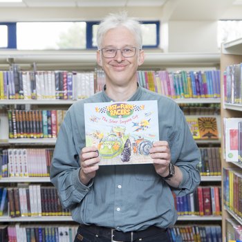An author wearing a grey shirt stands in an isle of books and holds a copy of his book