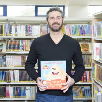 An author wearing a black shirt stands in an isle of books and holds a copy of his book