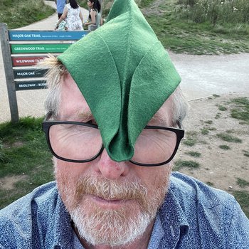 Photograph of Alistair Morrison wearing a green felt Robin Hood hat in front of Sherwood Forest signposts