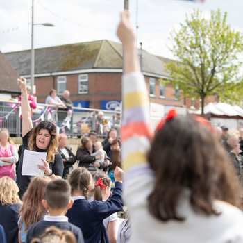 Children taking part in a singing activity with a young girl waving a flag in the foreground.