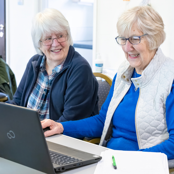 Two older women sat smiling whilst working on a laptop