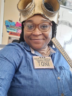 Investigator Toni smiling, wearing blue overalls with name badge, glasses and goggles on head
