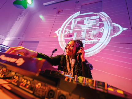A girl stands at a DJ set with strobe lights behind