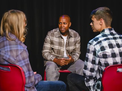 Three people wearing checked shirts sitting on red chairs talking.
