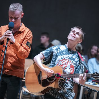 One male singing into a mic and one male playing guitar