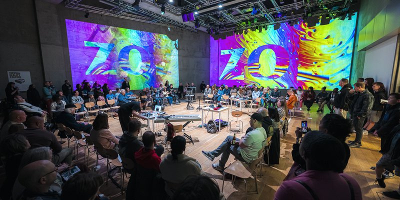 A photo from the Unlocked 2025 event at Nottingham Contemporary. Photo is taken from behind the audience awith a view of the wohle room inlcuding the audience stage area and digital graphics on the walls of the room.