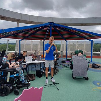 A boy in a blue T shirt centre stage singing into a microphone. Performing outdoors under a red, green and blue pergola. The floor is green