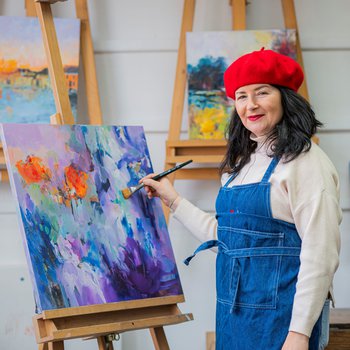 Alexandra is a white middle-aged woman with dark brown shoulder-length hair. She is wearing a red beret and blue artist's overalls and she's standing next to a painting.