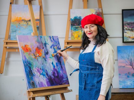 An artist in a blue denim apron stands in her studio next to an easel