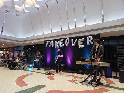 Musicians playing inside a shopping centre in front of balloons that read Takeover