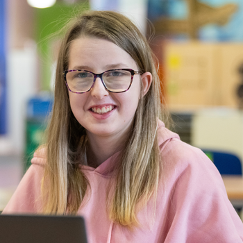 a young student with glasses smiles from behind a laptop