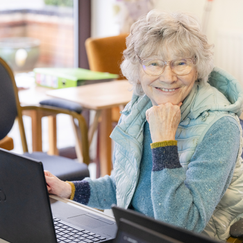 A very smiley older woman with short curly grey hair works at a laptop