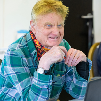 An older man with short red hair and bright blue eyes is smiling whilst working on a laptop