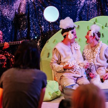 Two performers dressed as sheep, with white woolly costumes and curly headpieces, are kneeling and looking at each other, while another performer is playing guitar on the side.