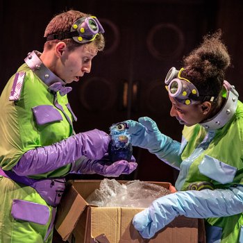 Two actors dressed for outer space looking in a cardboard box.