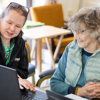 An older white woman with short hair is smiling whilst a tutor is helping her to use a laptop. The tutor is a middle-aged white woman with reddish-brown hair tied back in a ponytail.