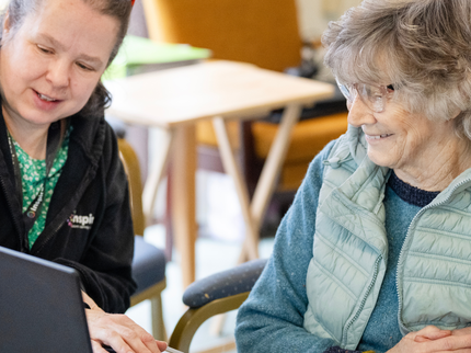 An older white woman with short hair is smiling whilst a tutor is helping her to use a laptop. The tutor is a middle-aged white woman with reddish-brown hair tied back in a ponytail.