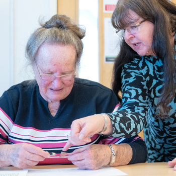 Learner wearing a red and black top is receiving help from tutor wearing a blue and black patterned top.