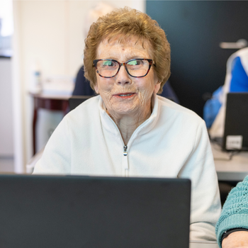 An older woman with short red hair and glasses is sat behind a laptop