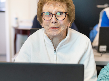 An older lady with short curly ginger hair is smiling from behind a laptop