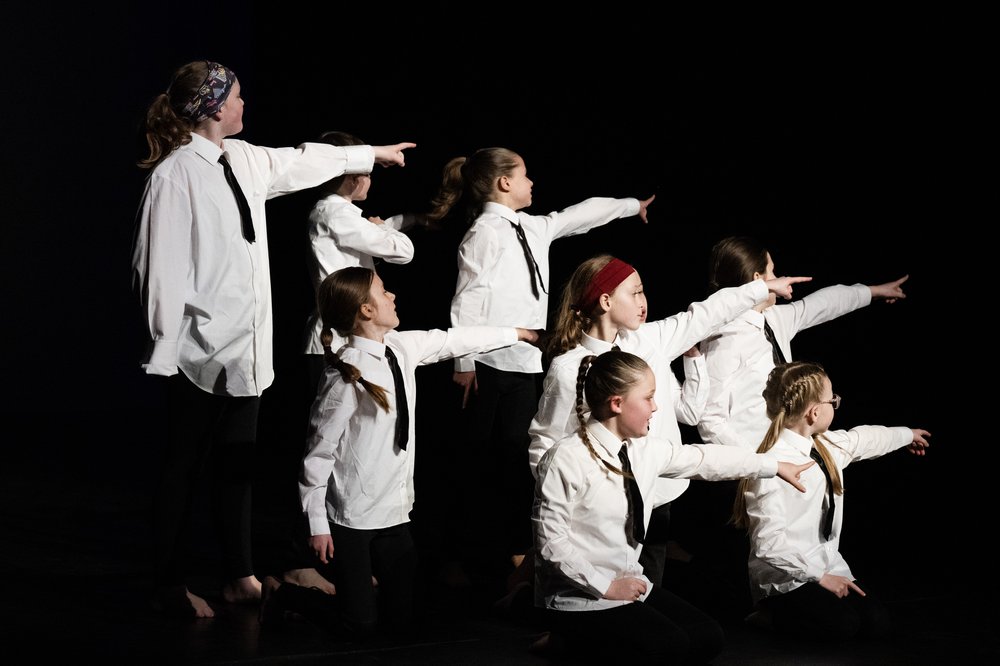 12 young dancers on a black stage, with their arms out at different angles. They are wearing black and gold costumes