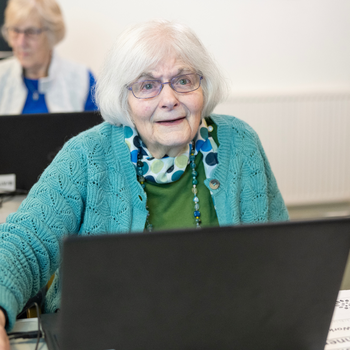 An older woman with a short white bob haircut and glasses smiles as she uses a laptop