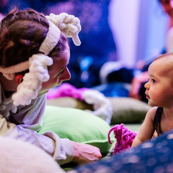 A performer dressed as a sheep, with a white woolly costume and curly headpiece, low to the ground and interacting with a baby who is looking back at the performer.
