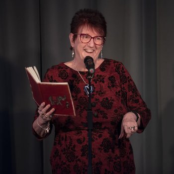 Lady on stage holding a book and reciting into a microphone