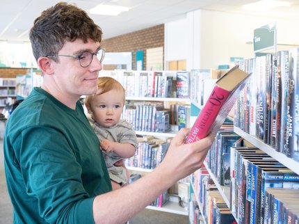 A man looking selecting a library book, holding his baby