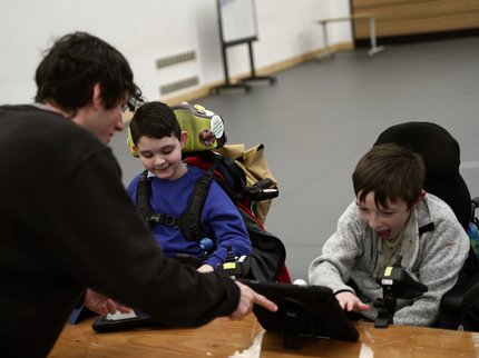 A male music tutor sharing a digital tablet with two young boys, who are smiling and engaged as they look at the screen together.