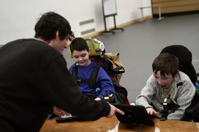 A male music tutor sharing a digital tablet with two young boys, who are smiling and engaged as they look at the screen together.