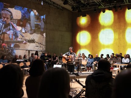 Image with a male in the centre holding a guitar and singing. With yellow round lights in the background, audience sat all around
