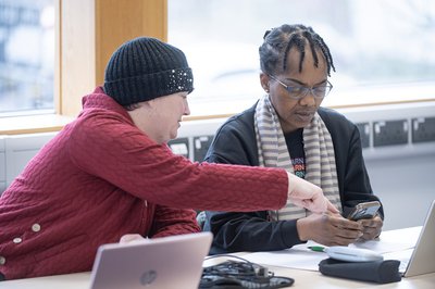 Two learners sit with laptops at a table in their local library.