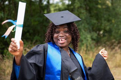 A lady wears a graduation cap and gown, smiling and celebrating with a scroll in her hand.