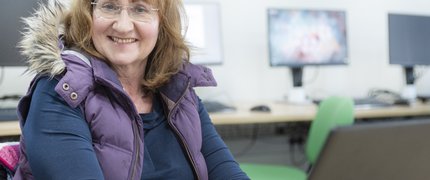 A middle aged woman with light brown shoulder length hair wearing glasses, a purple gilet and navy blue long sleeve top sits at a computer and smiles.