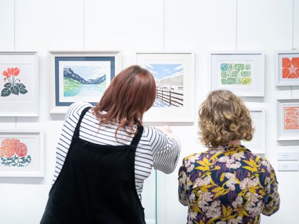 Two women are facing away from the camera whilst they look at a display of art as part of an exhibition.