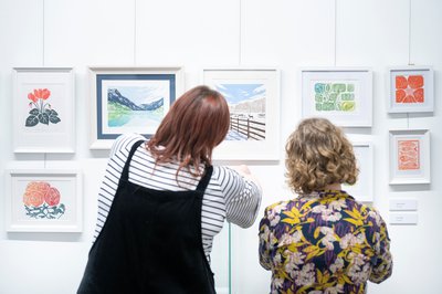 Two women are facing away from the camera whilst they look at a display of art as part of an exhibition.