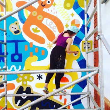 Artist Alex stood on scaffolding painting a snake, part of a large wall mural in blue, yellow and orange colours