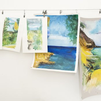 Six finished water colour paintings are pegged to a line in an artists studio. They show beach scenes.