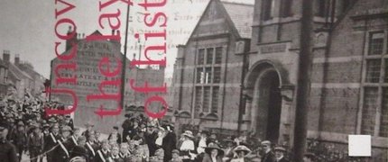 Black and white photo of school children in uniform marching in front of old school